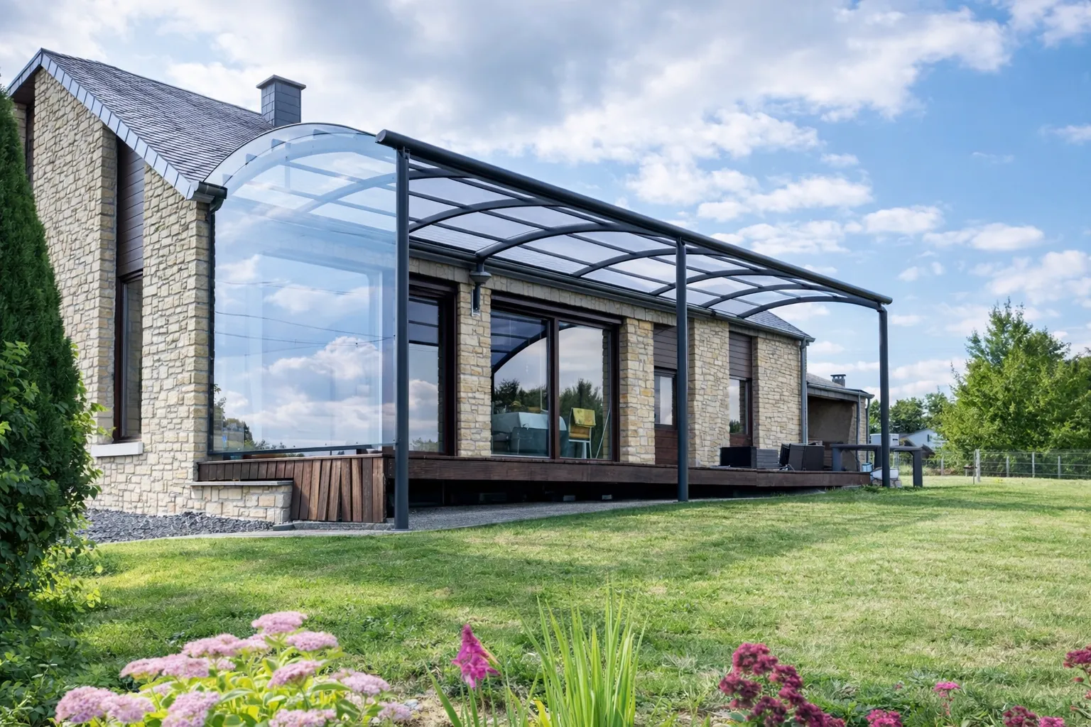 Pergola Ovalux aluminium adossée à une maison en pierre, toiture polycarbonate cintrée transparente sur terrasse bois et jardin